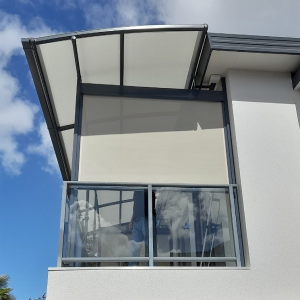 A modern grey retractable awning extends over a patio area of a light-coloured house with a dark tiled roof. Green lawn and shrubs surround the patio, and large bifold doors are visible under the awning.