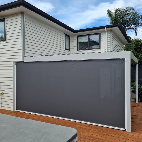 A modern grey retractable awning extends over a patio area of a light-coloured house with a dark tiled roof. Green lawn and shrubs surround the patio, and large bifold doors are visible under the awning.